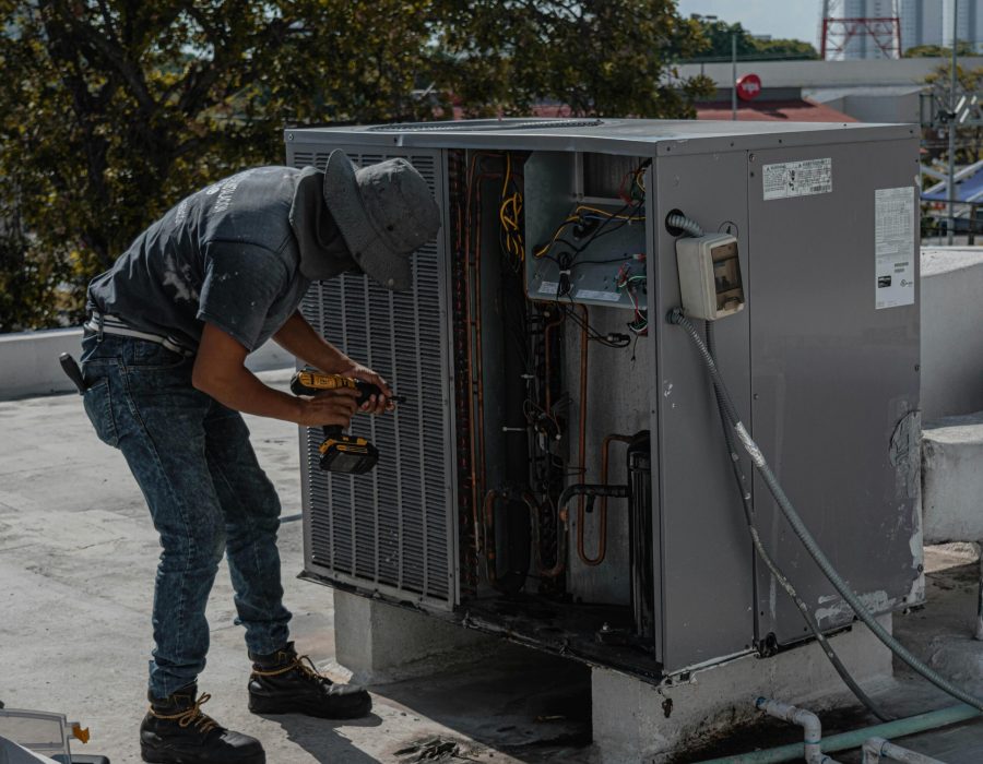 A worker in a bucket hat repairs an outdoor air conditioning unit on a rooftop.